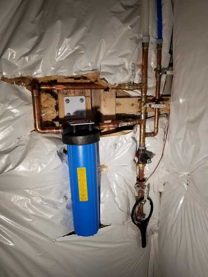 Blue water filter with exposed copper pipes and valves against a plastic-covered wall