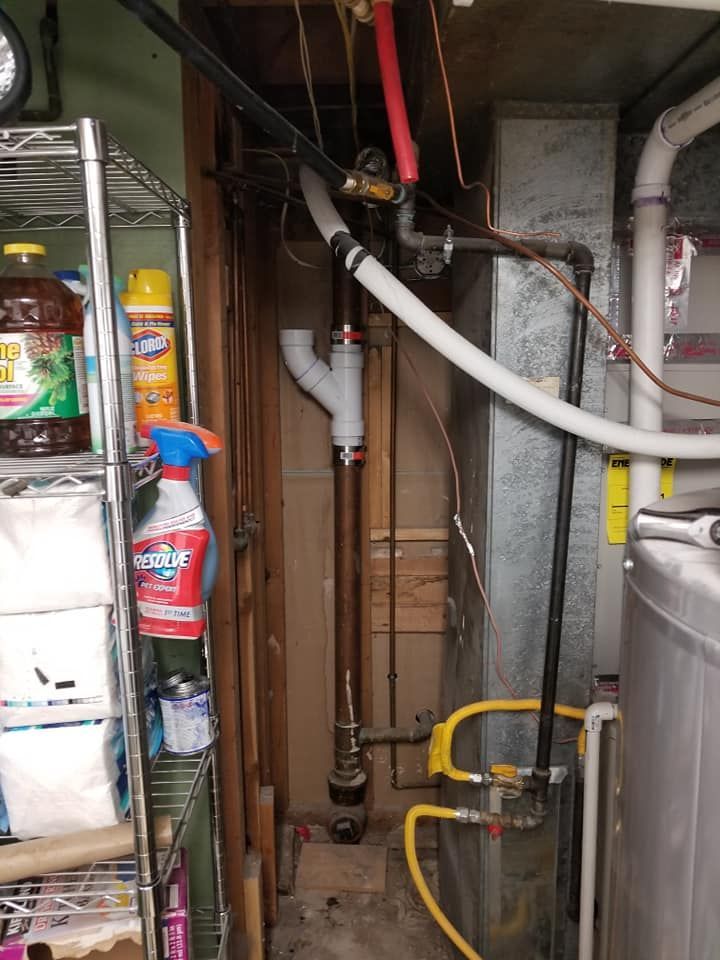 Cluttered utility closet with exposed pipes, hoses, and cleaning supplies on shelves.