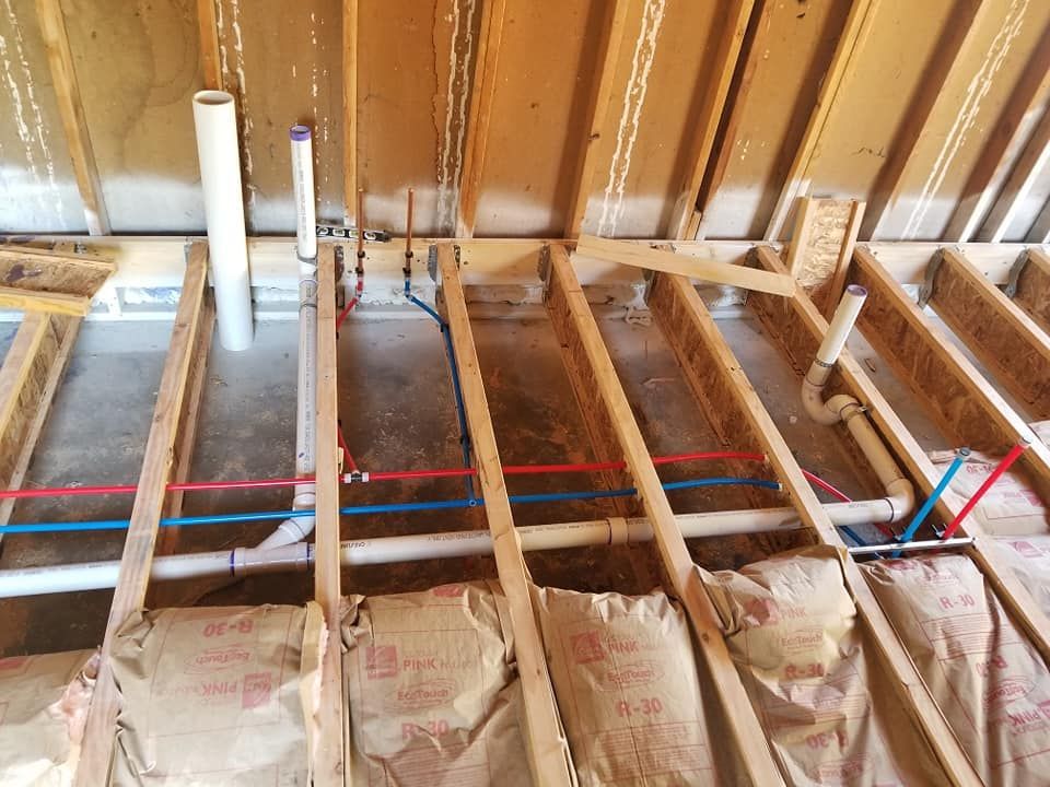 Exposed attic framing with insulation, white PVC pipes, and red and blue electrical lines running across joists