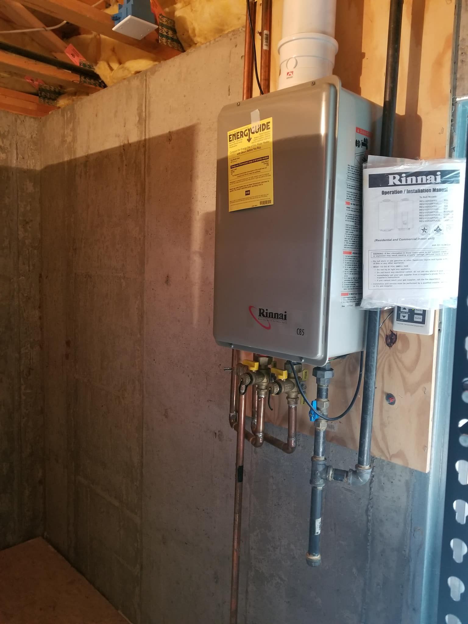 Wall-mounted gray tankless water heater with pipes and a paper notice in a basement utility area