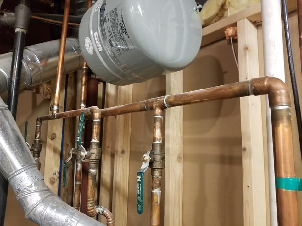 Copper pipes and valves beneath a gray expansion tank in a basement utility area
