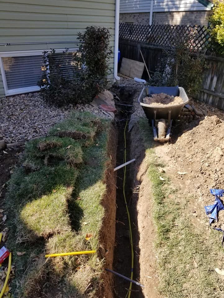 Trench dug between lawn edges beside a house, with a wheelbarrow and garden tools nearby.