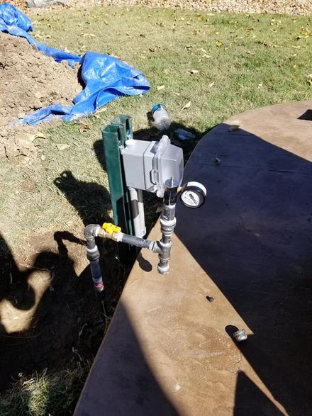 Water meter and pipes mounted beside a wooden board outdoors on grass, with a blue tarp nearby