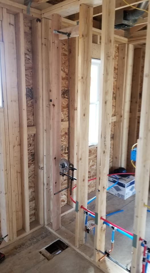 Unfinished house framing with exposed wooden studs, plumbing pipes, and a floor drain opening.