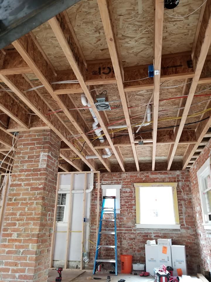Renovation site with exposed wooden ceiling joists, brick walls, ladder, and tools in a room under construction.