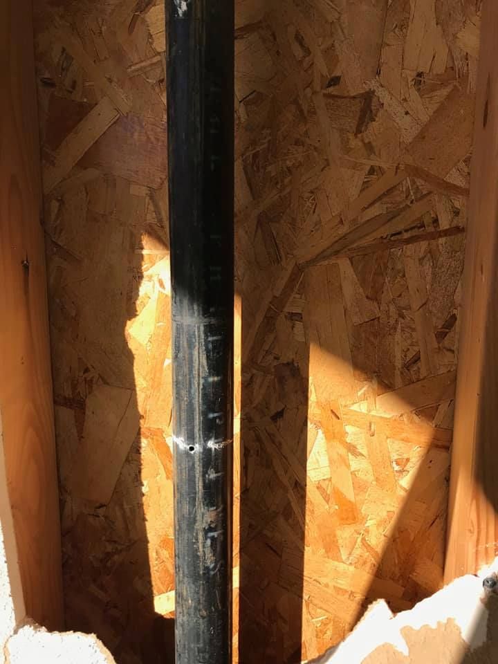 Black vertical pipe centered in a plywood-framed wall opening with sunlight shining through.