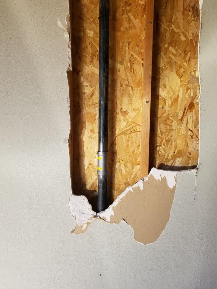 Damaged drywall exposing wooden studs and black pipe inside a wall.