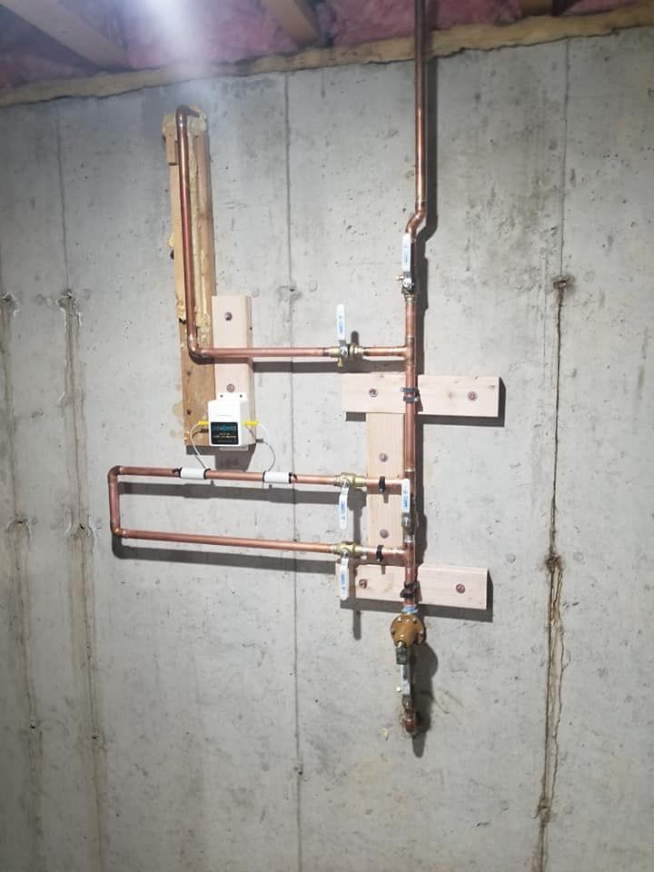 Copper and PVC pipes mounted on a concrete basement wall, with a valve and meter assembly.