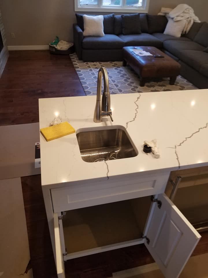 Modern kitchen island with sink and open cabinet, overlooking a living room with gray sectional sofa.