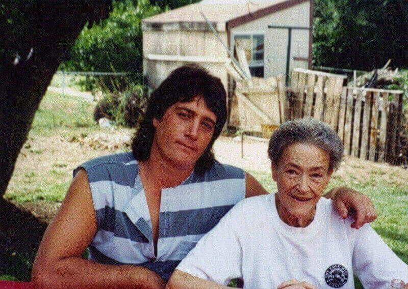 Two people sitting outdoors, one with an arm around the other, in front of a house and fence.