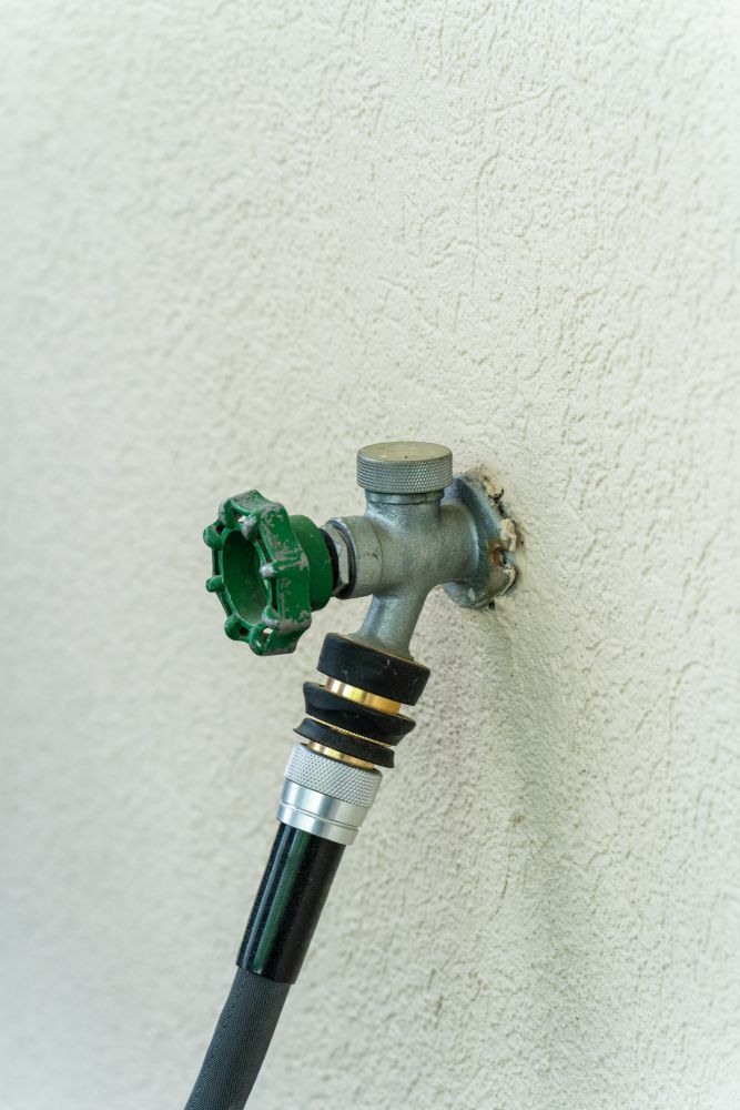Green-handled garden hose spigot mounted on a stucco wall with a hose attached.