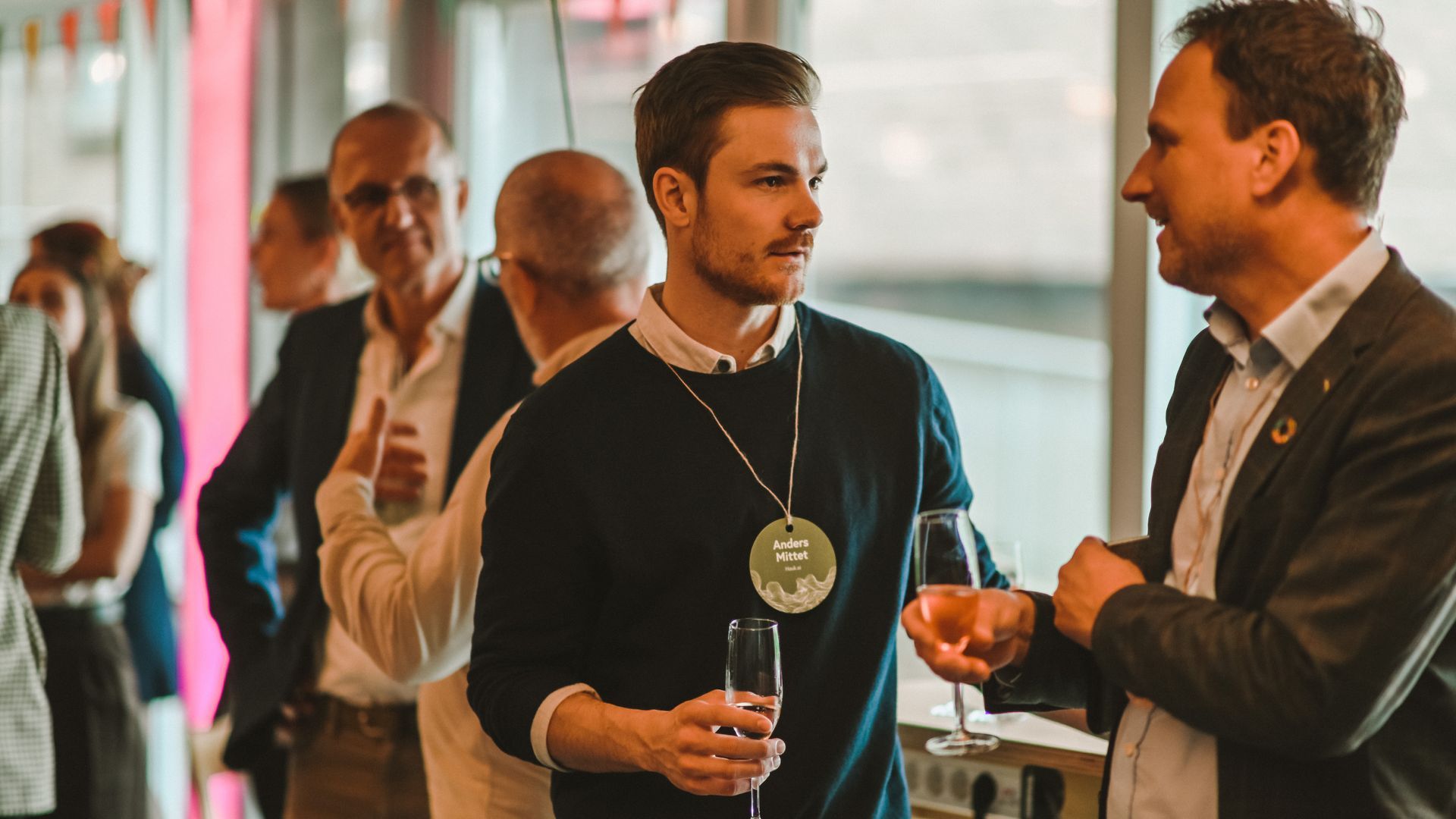 Two men chatting at an event, one holding a drink, wearing a lanyard; other people in background.