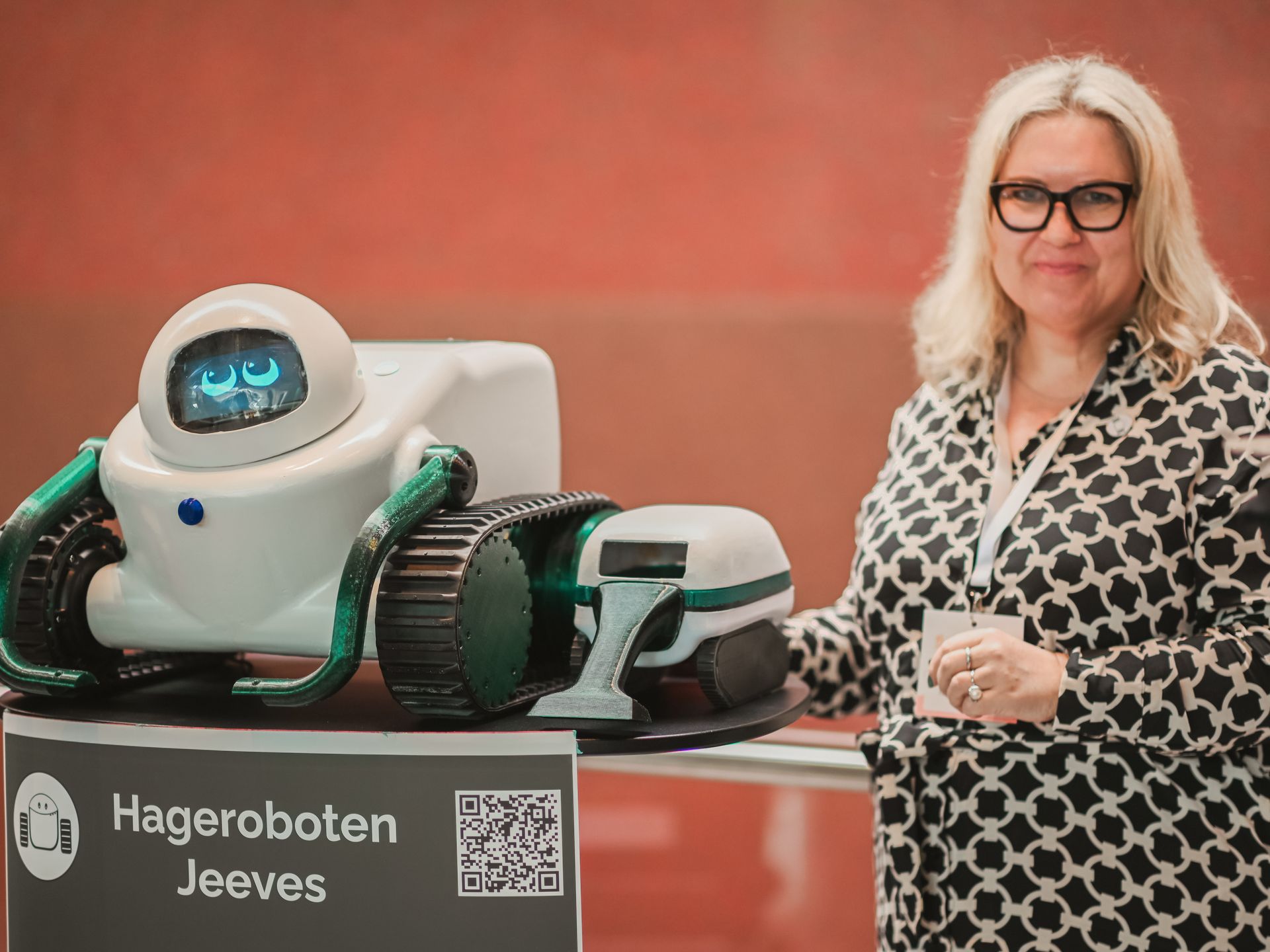 Woman next to a white robot named Jeeves; both stand by a sign with text and a QR code.