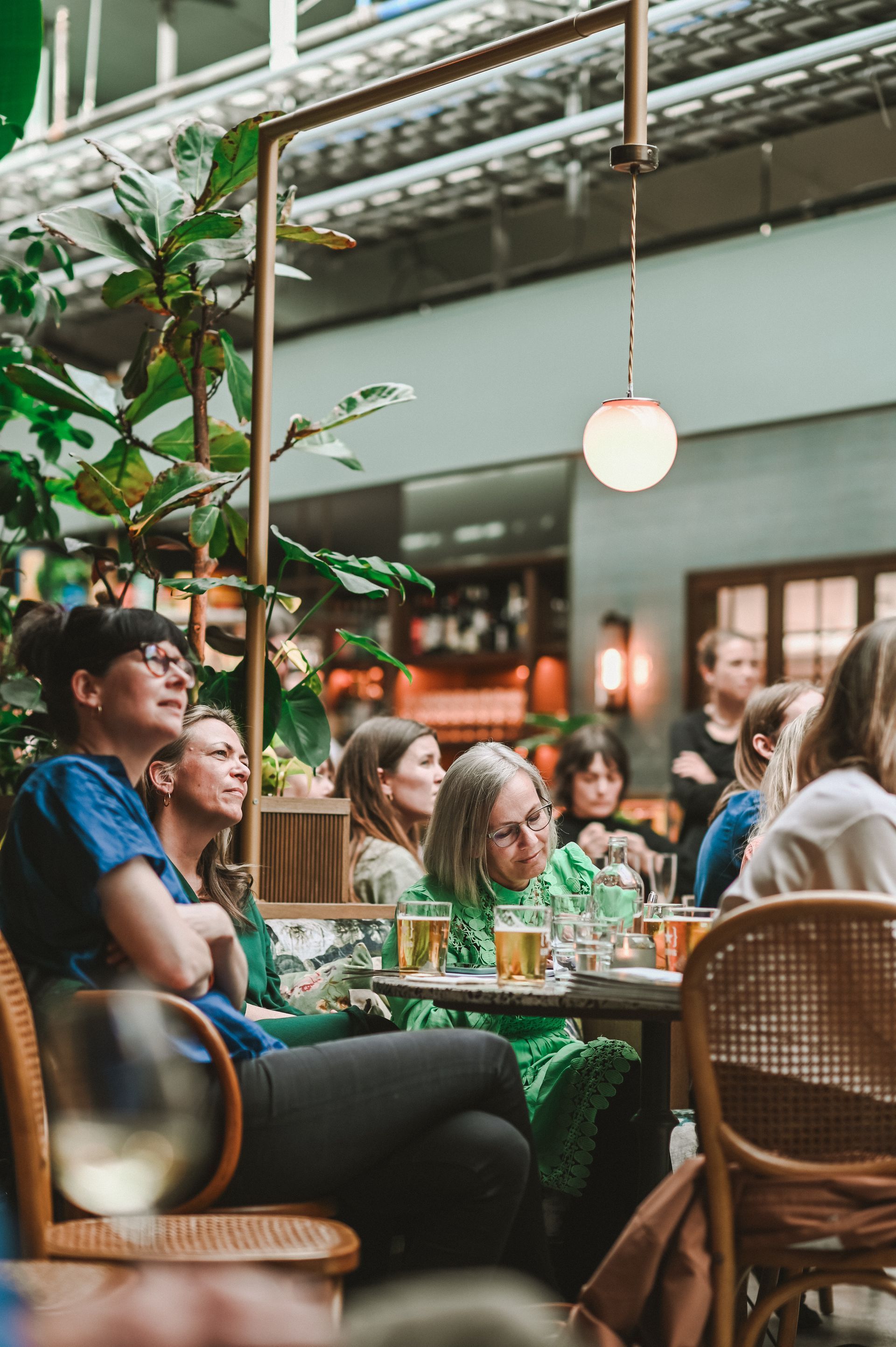 People seated at outdoor tables, drinking and watching something. Greenery and a hanging light fixture.