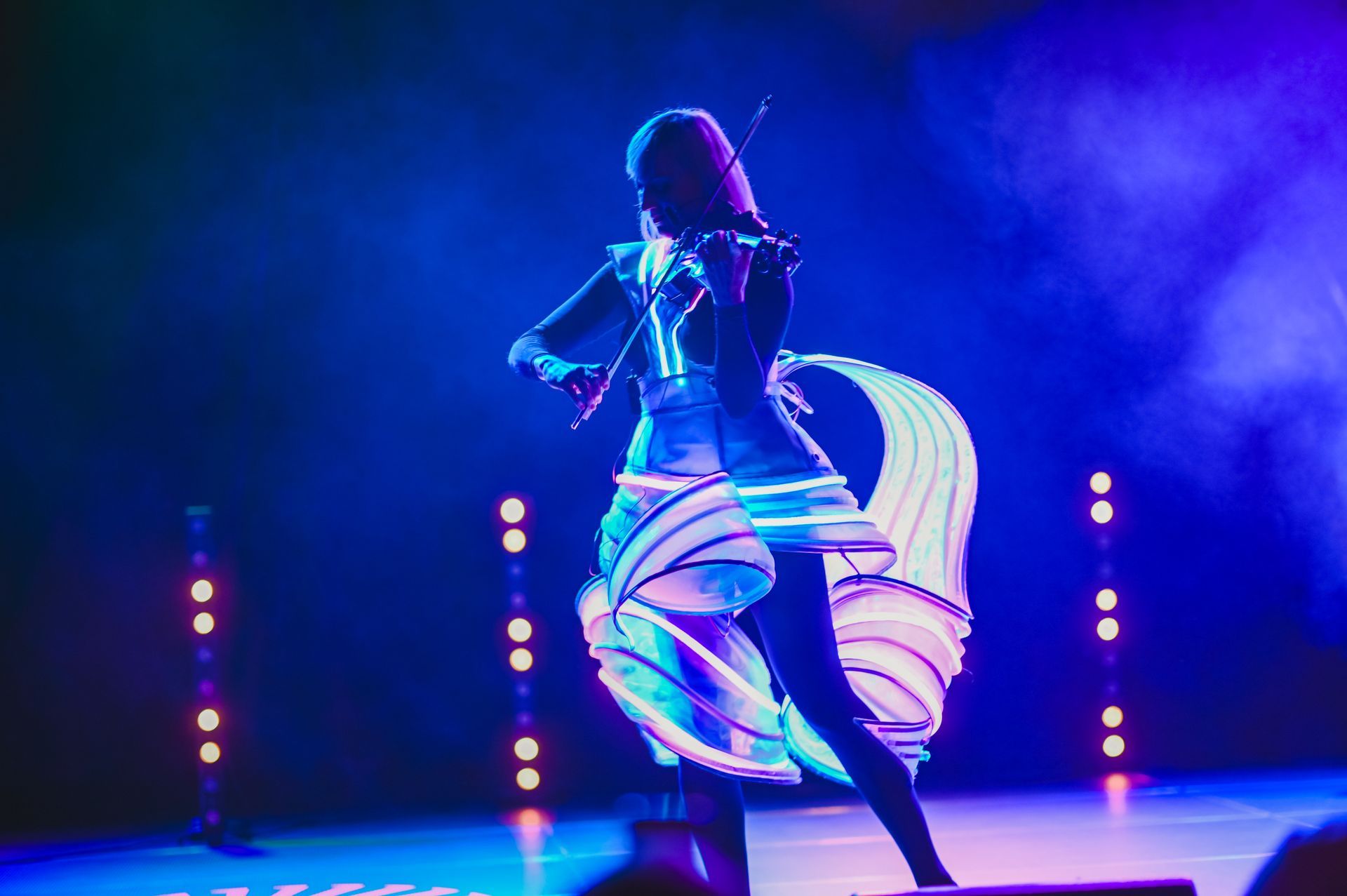 Woman in illuminated costume plays violin on stage with colorful lights.