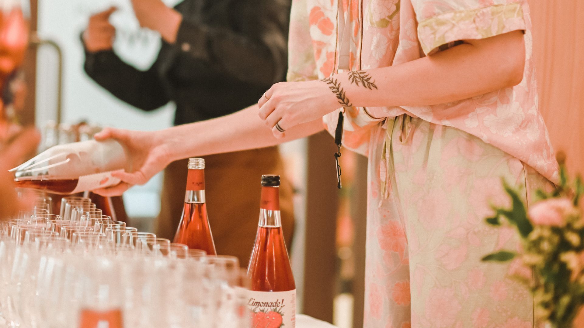 Person pouring drink into glasses at a gathering, pink-toned, with bottles and floral accents.