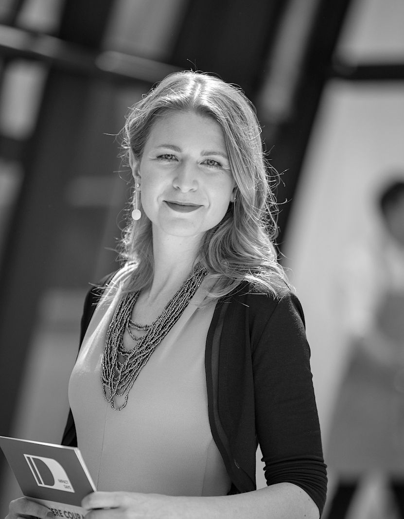 Woman in light top and dark cardigan, beaded necklace, smiling, holding brochure in front of a building.
