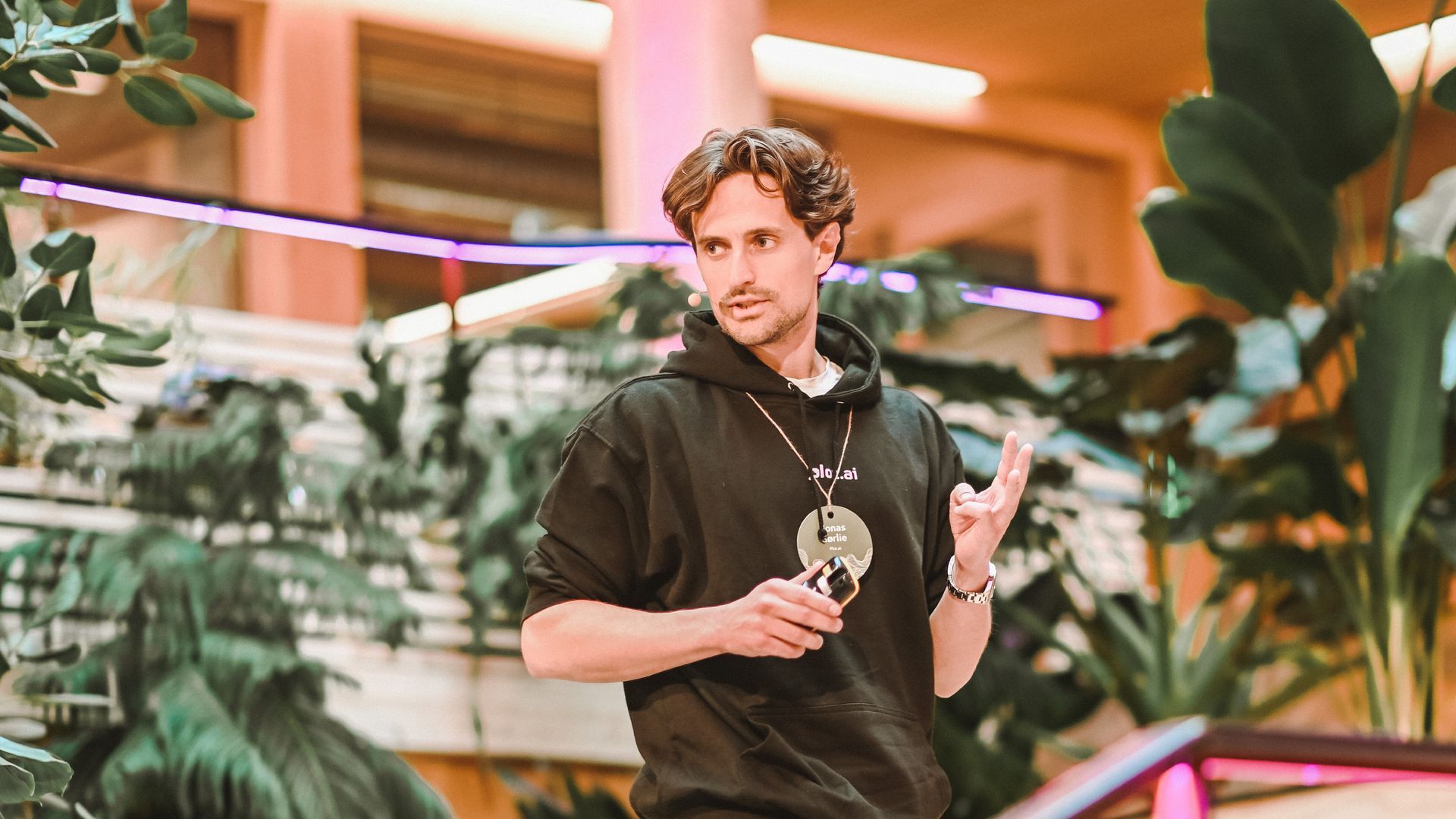 Man in a black hoodie gesturing with two fingers, surrounded by plants, speaking at a venue.