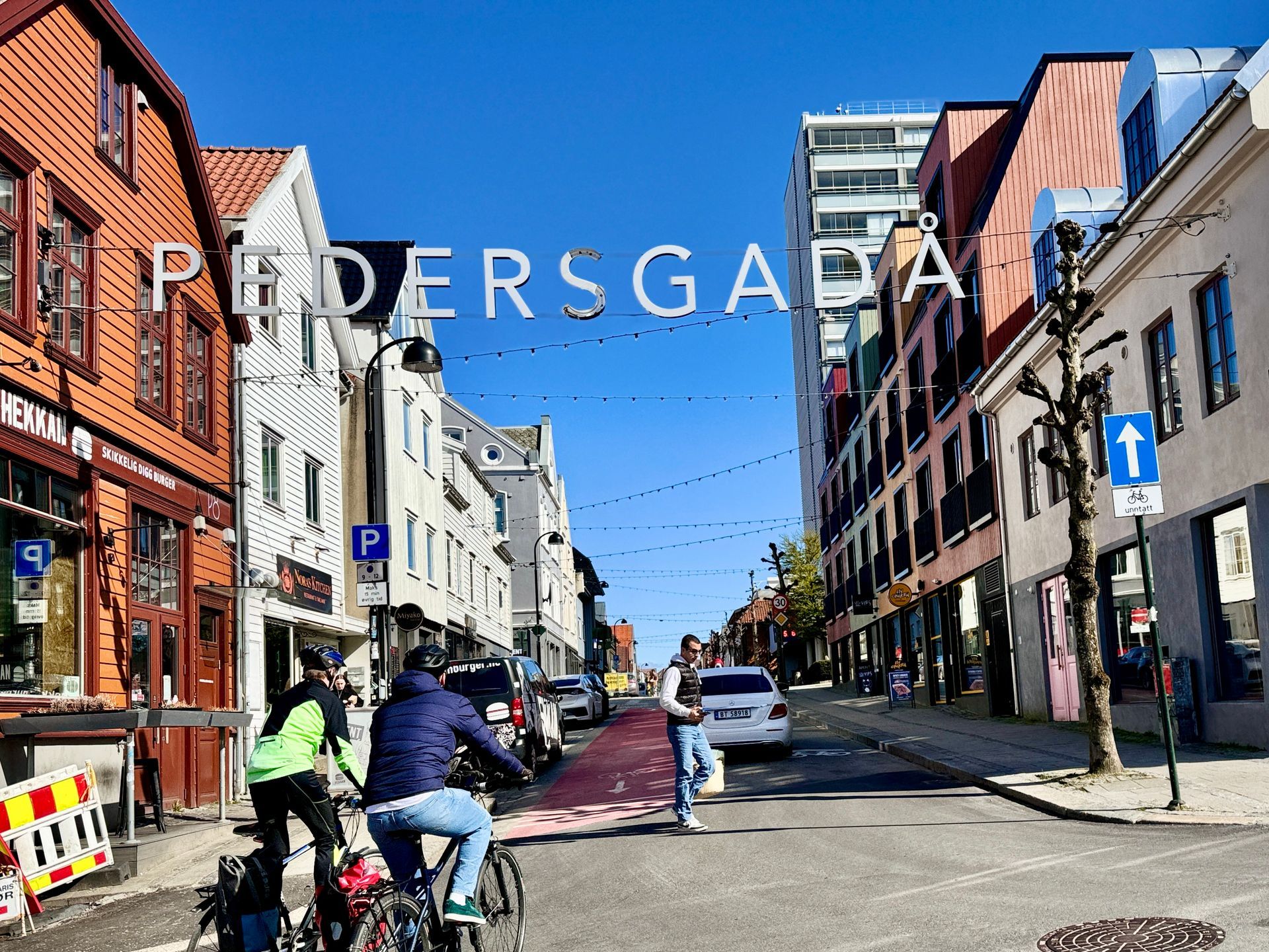Street scene in Pedersgata, Norway, with buildings, street sign, cyclists, pedestrians, and parked cars.