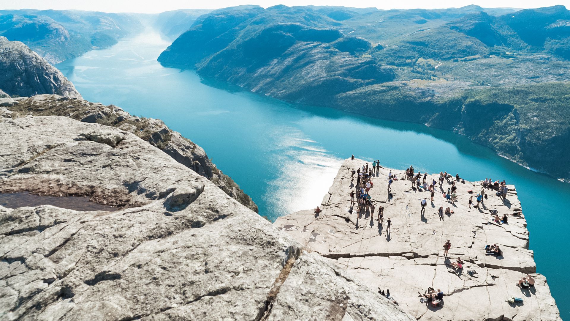 View of Preikestolen cliff overlooking a fjord with many people gathered on the rock plateau.