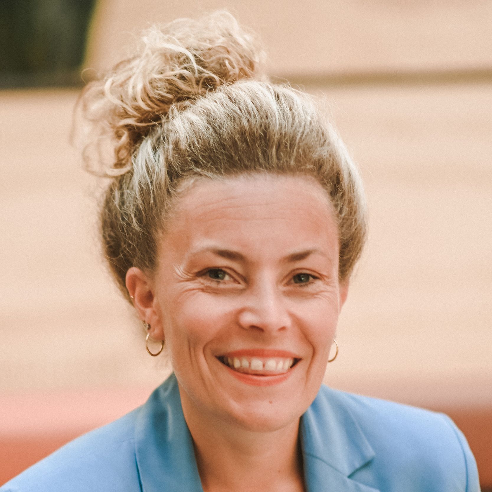 Woman with blonde hair in a bun, wearing a blue blazer, smiles at the camera. Outdoor setting.