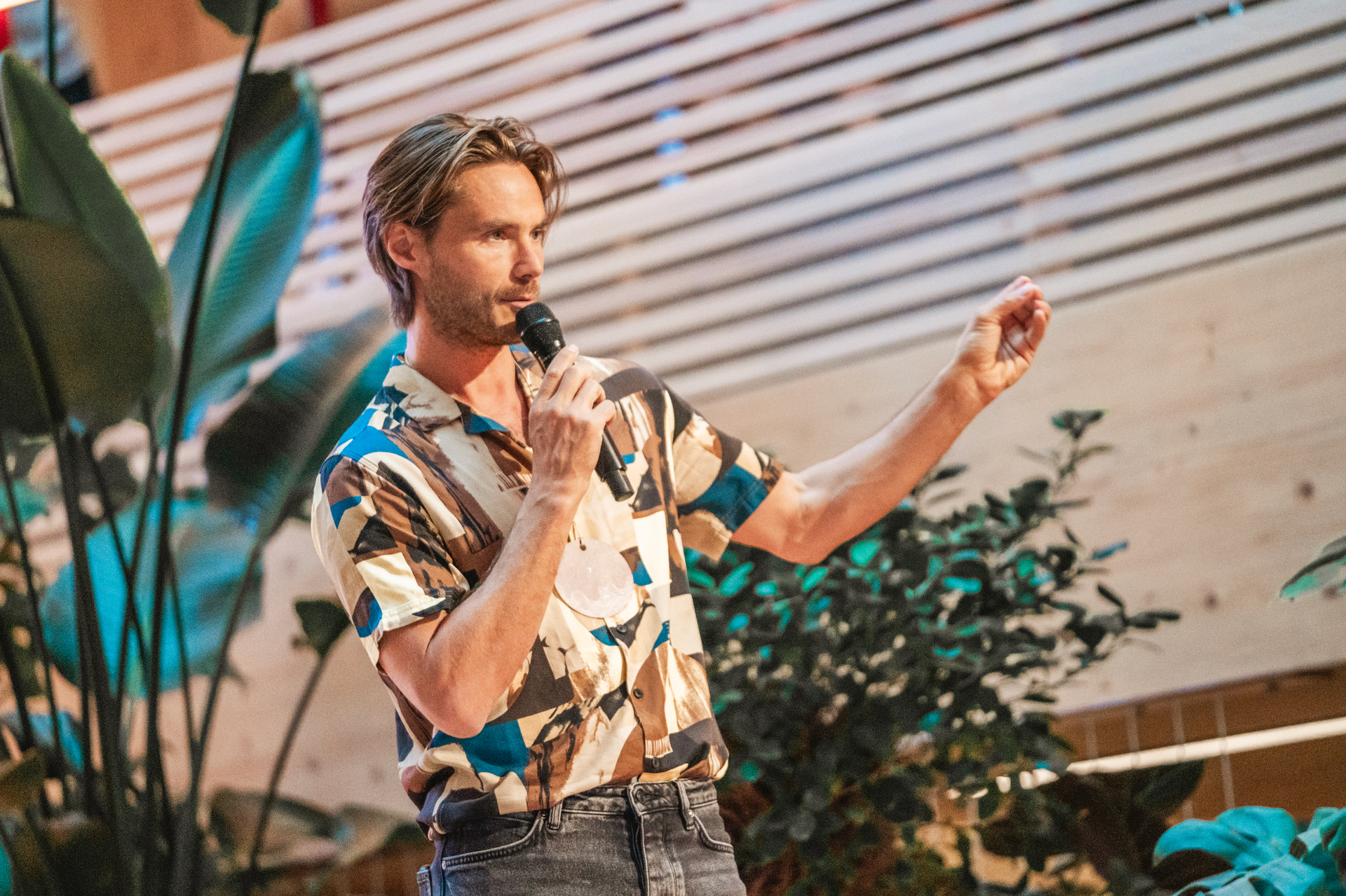 Man speaking into a microphone, gesturing with his right hand. He's in a room with plants and a wood panel backdrop.
