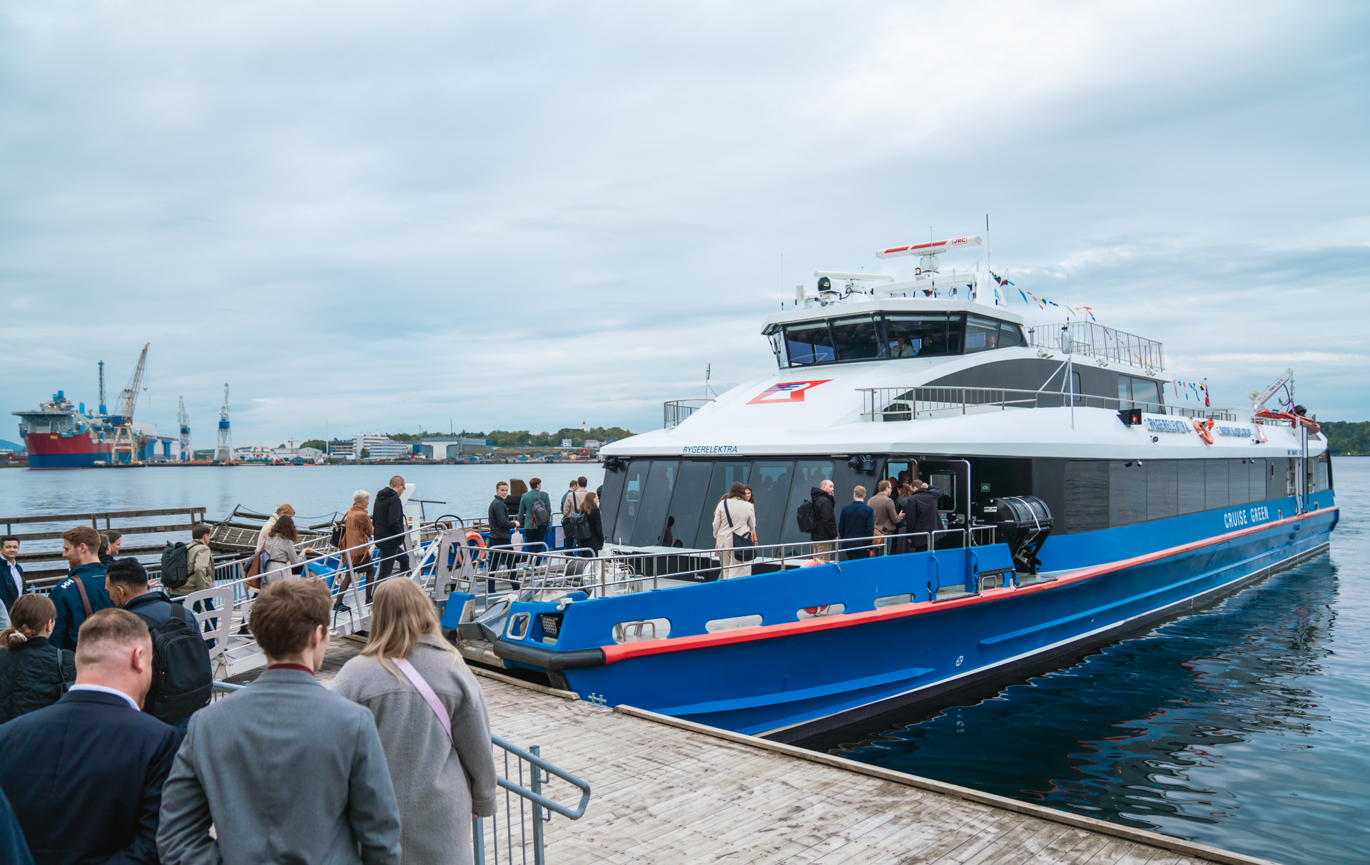 Blue ferry at a dock, people boarding. Harbor with cranes and overcast sky.