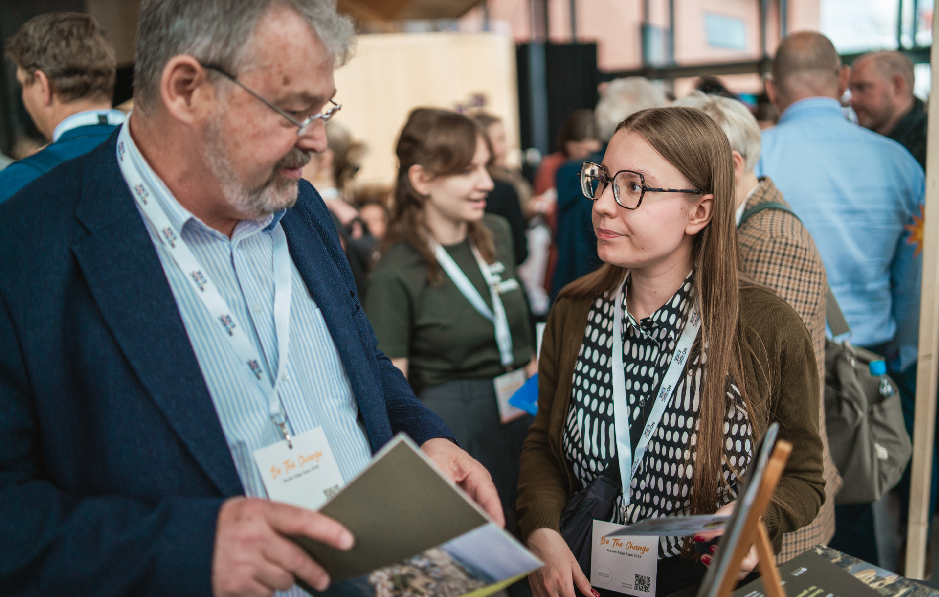 Man in suit talks to a woman with glasses, both wearing lanyards, at an event.