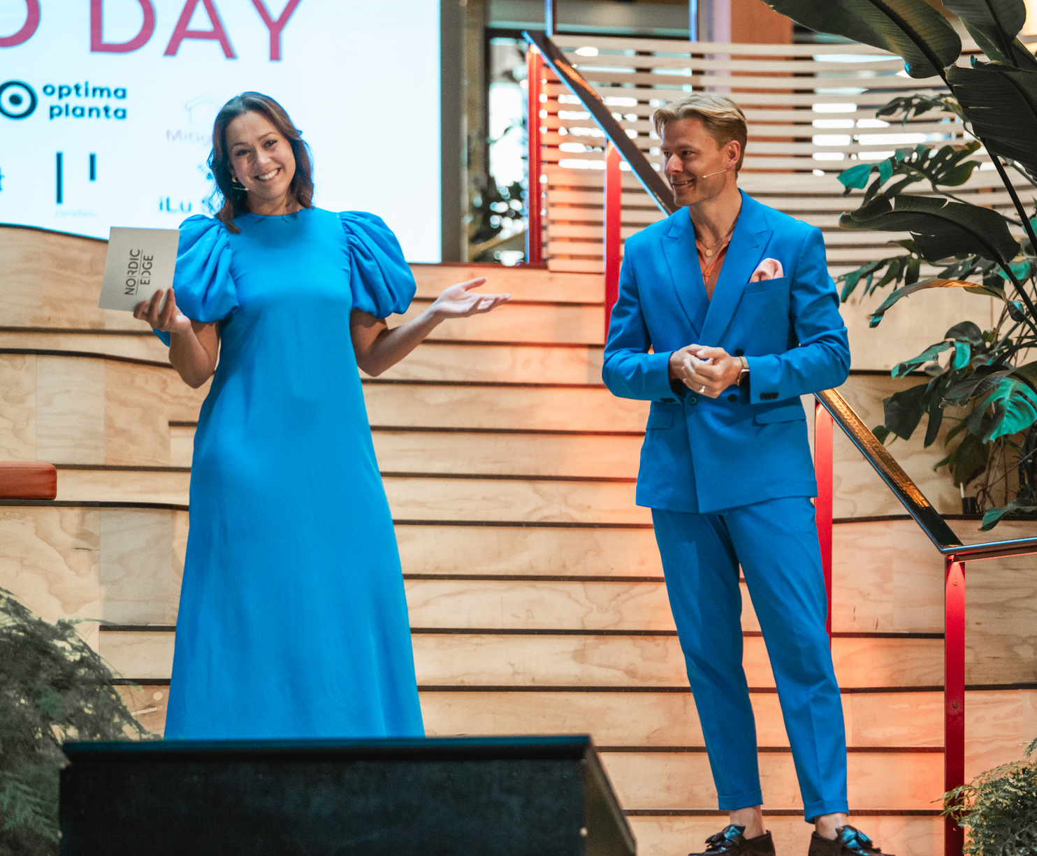 Woman in blue dress and man in blue suit on stage, smiling and presenting.