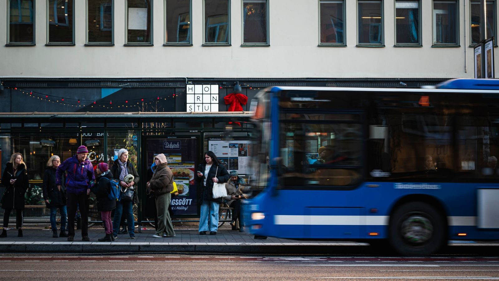 People waiting at a bus stop next to a blue bus. Building facade with windows in background.