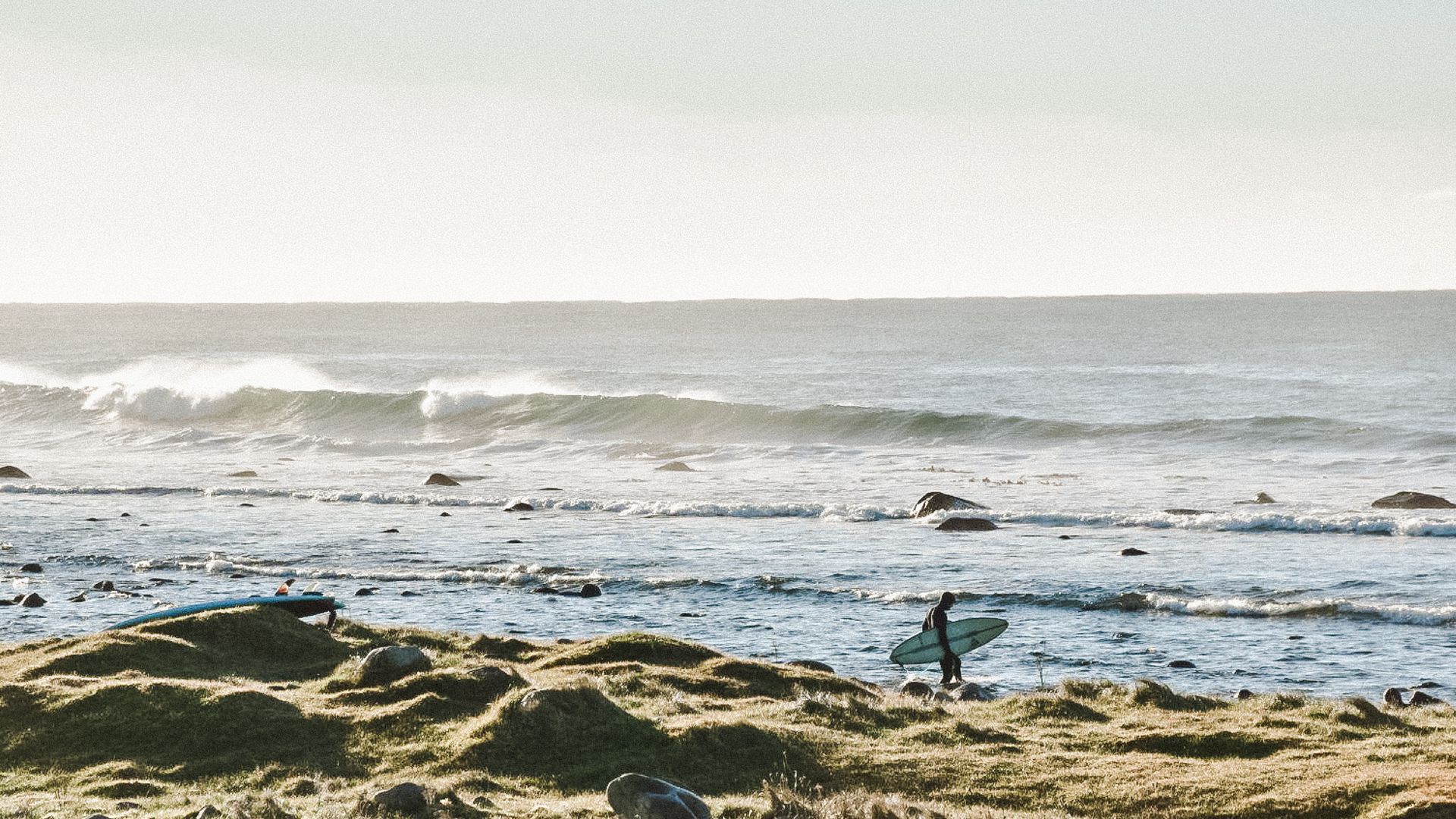 Surfer walks across grassy dunes towards the ocean, carrying a surfboard, with waves breaking in the background.