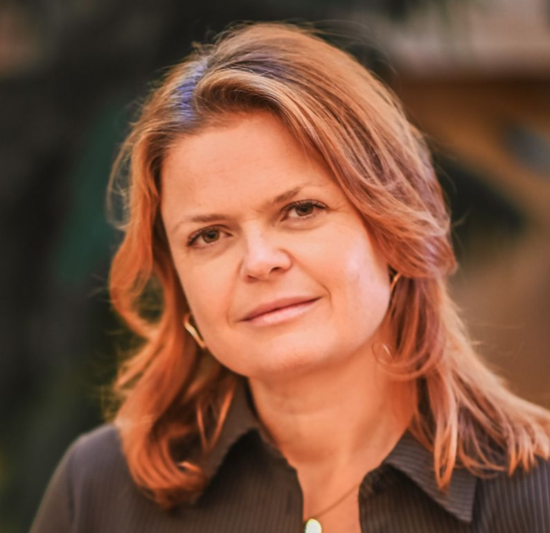 Woman with light brown hair smiles, wearing a cream turtleneck and dark blazer.