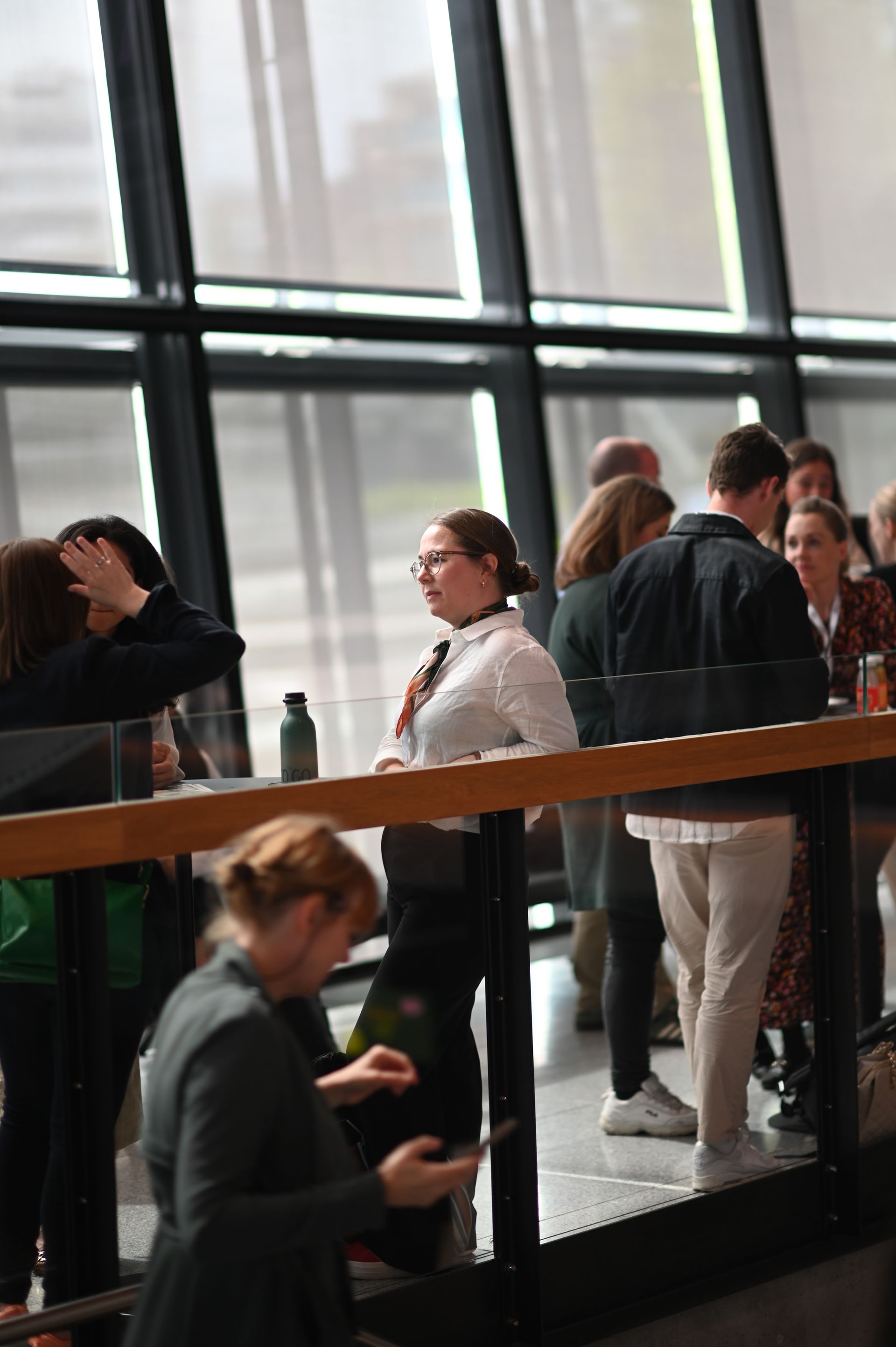People gathered near large windows, some talking, others looking. Building interior with a railing.