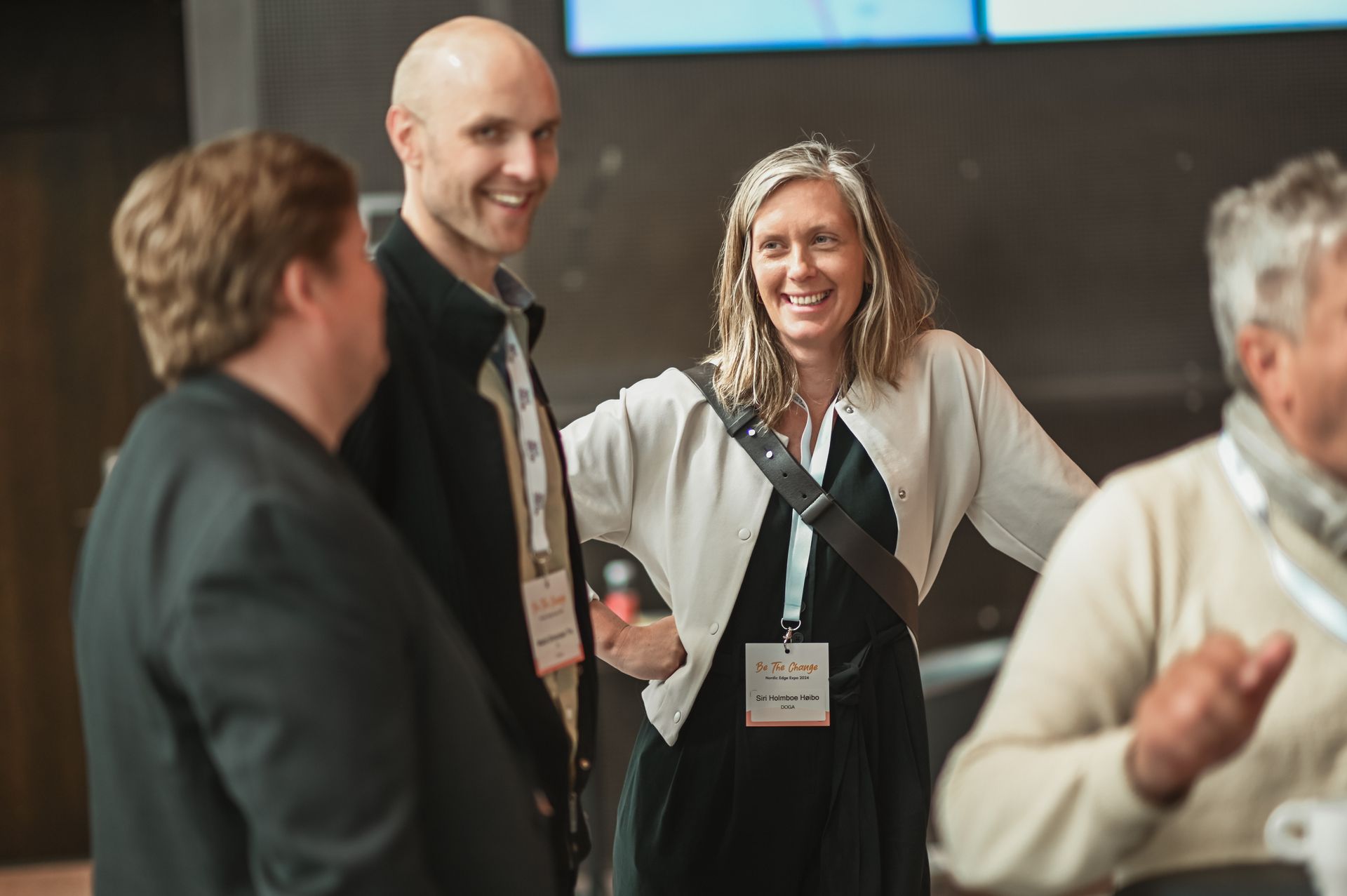 People in a group smiling and talking, some wearing lanyards, indoors.