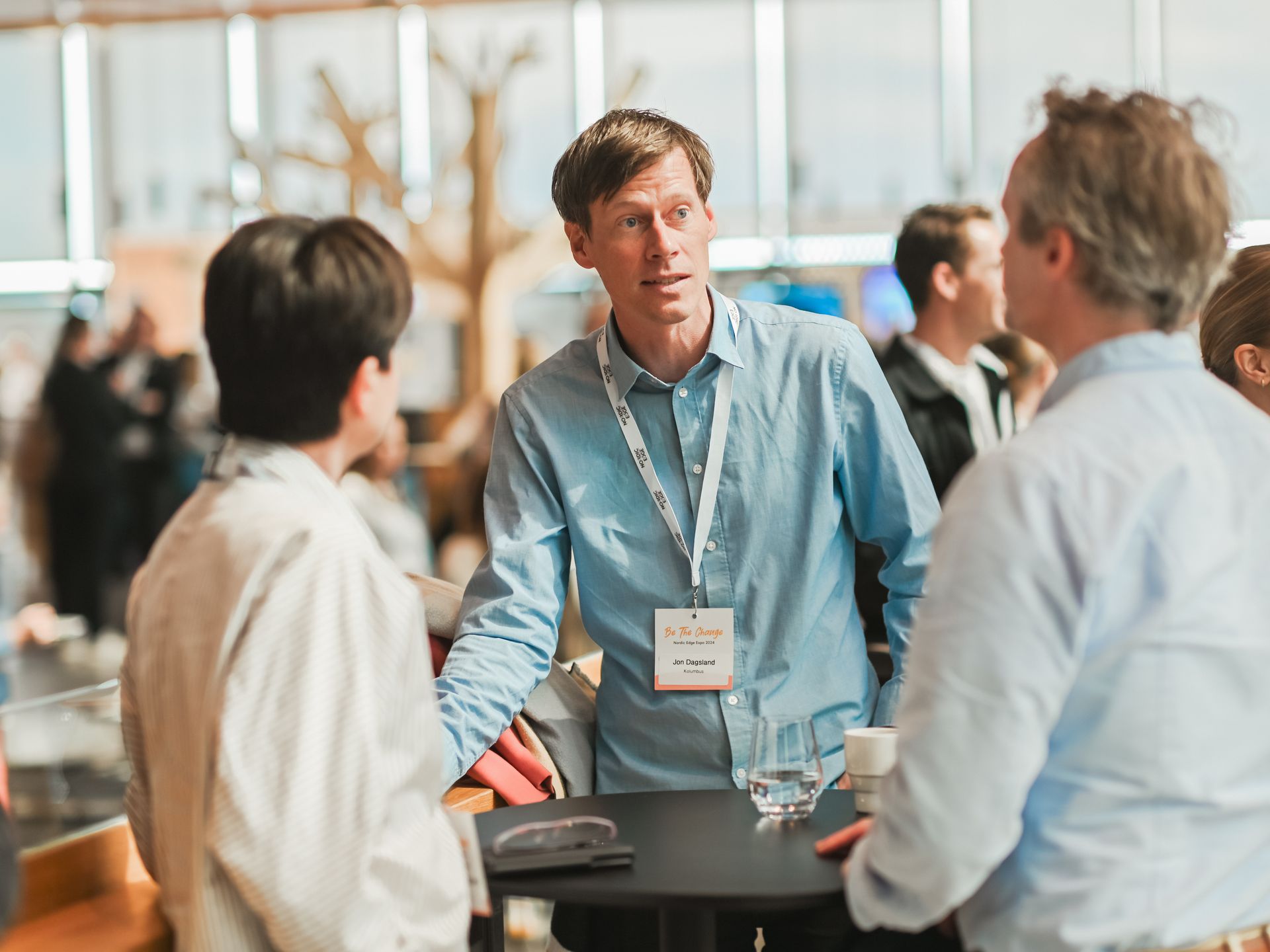 Three people at an event, conversing. Man in blue shirt speaks, others listen. Black table with items.