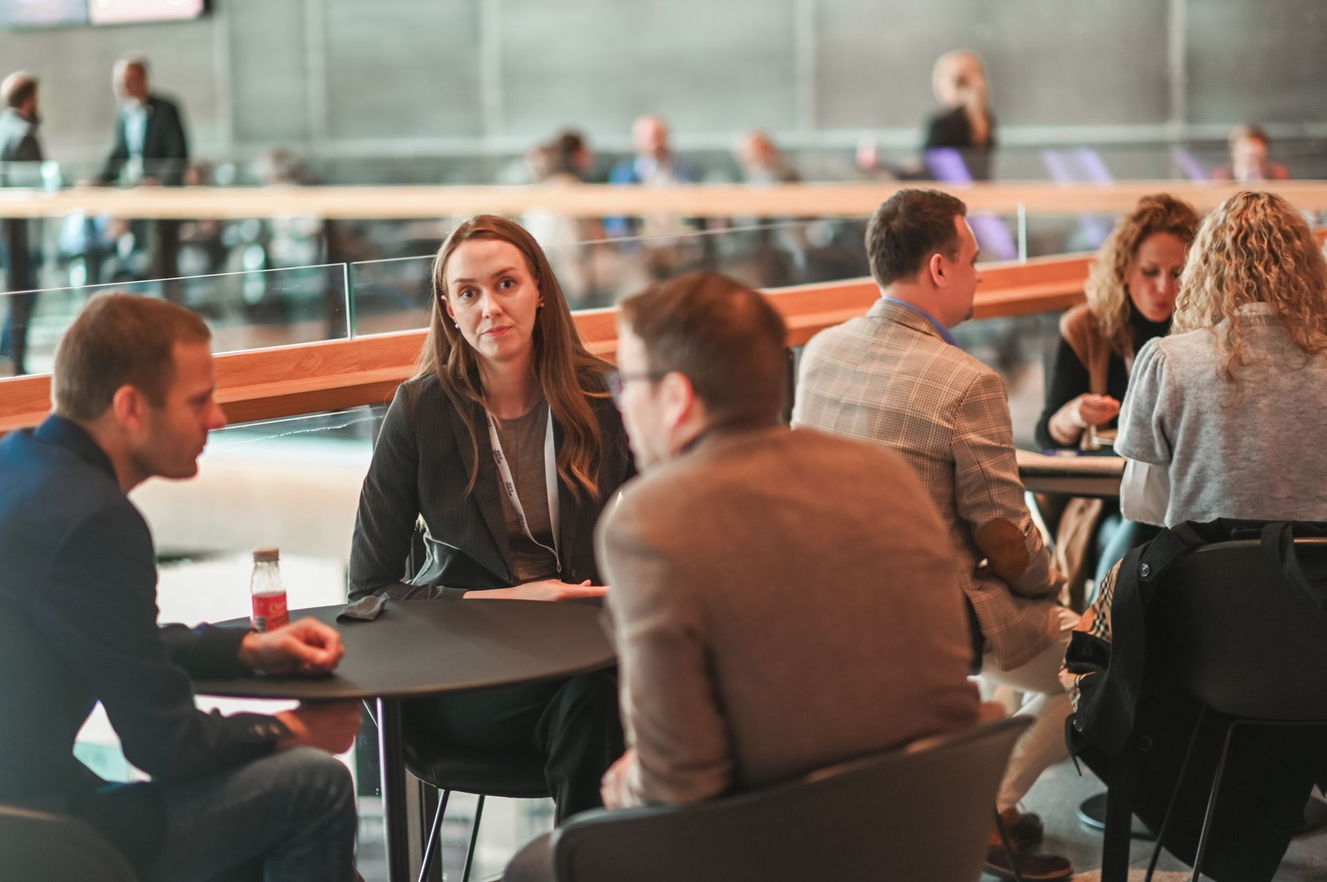 People networking at tables in a modern building. Some are talking, others are listening. Natural light illuminates the scene.