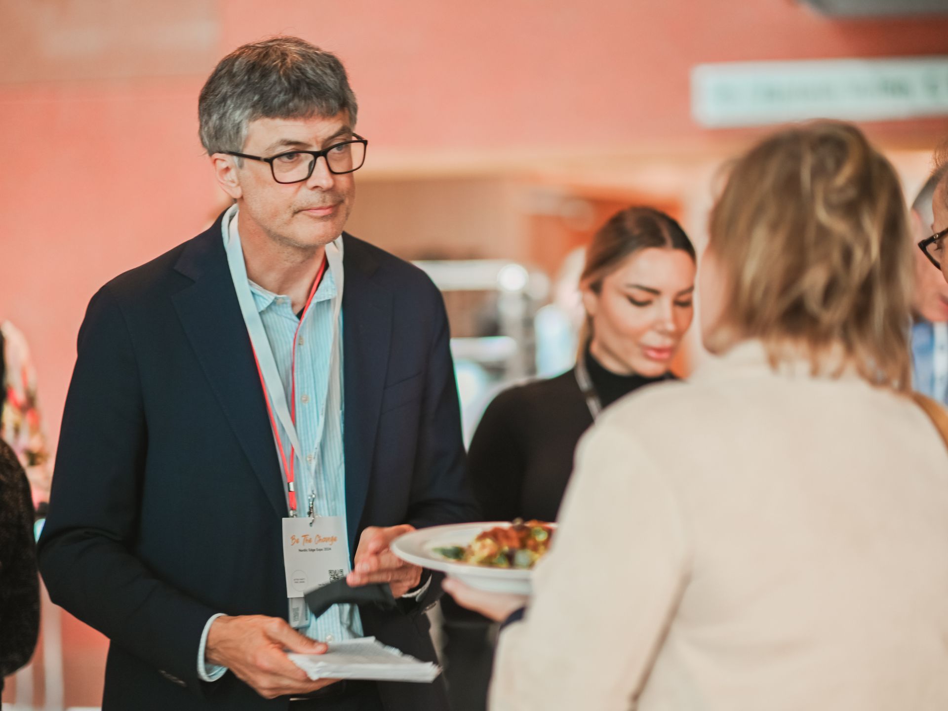 Man in glasses at an event, holding a plate. Others are nearby; warm-toned room.