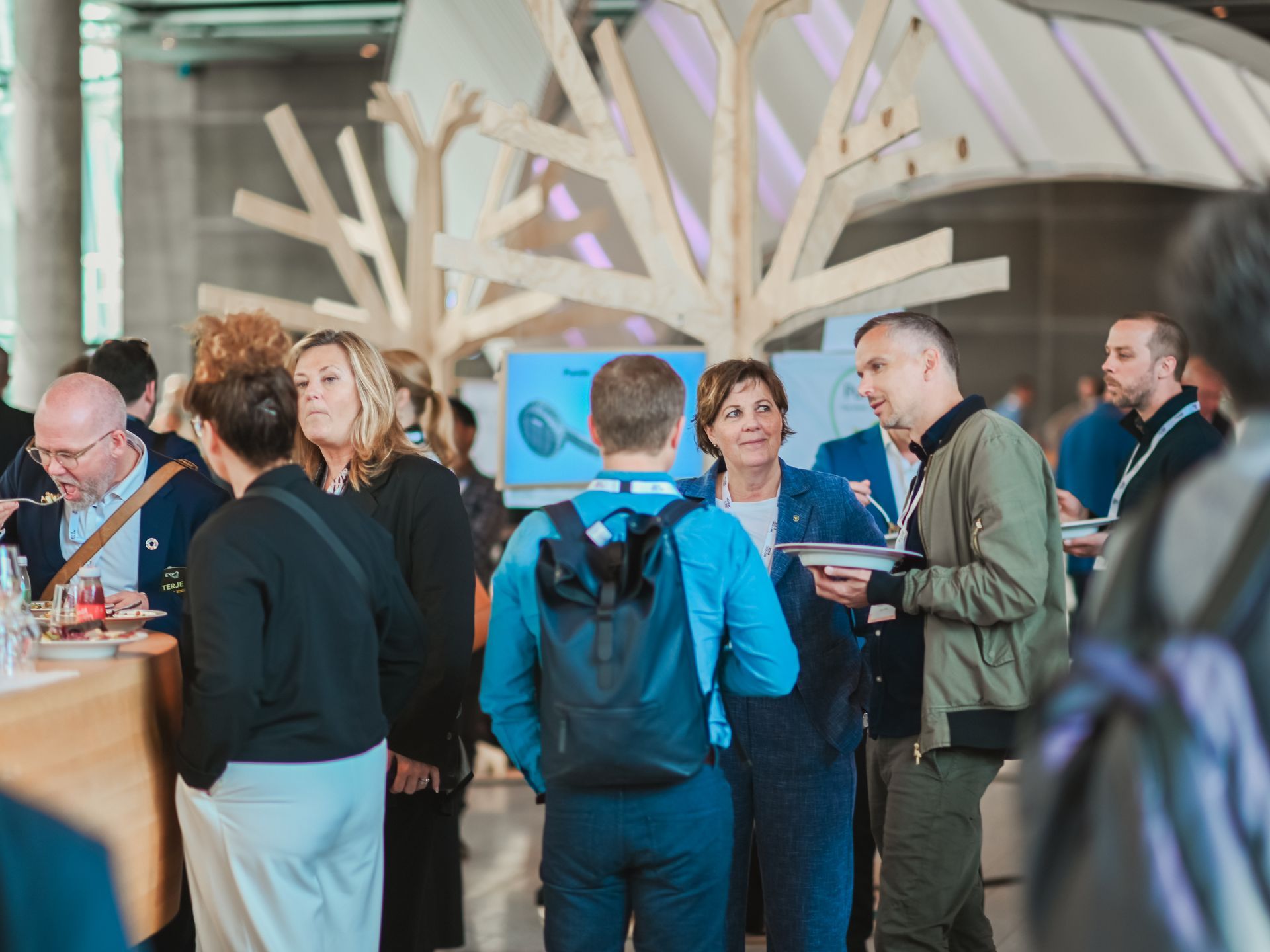 Attendees mingle at a conference, conversing and holding plates of food near wooden art installation.