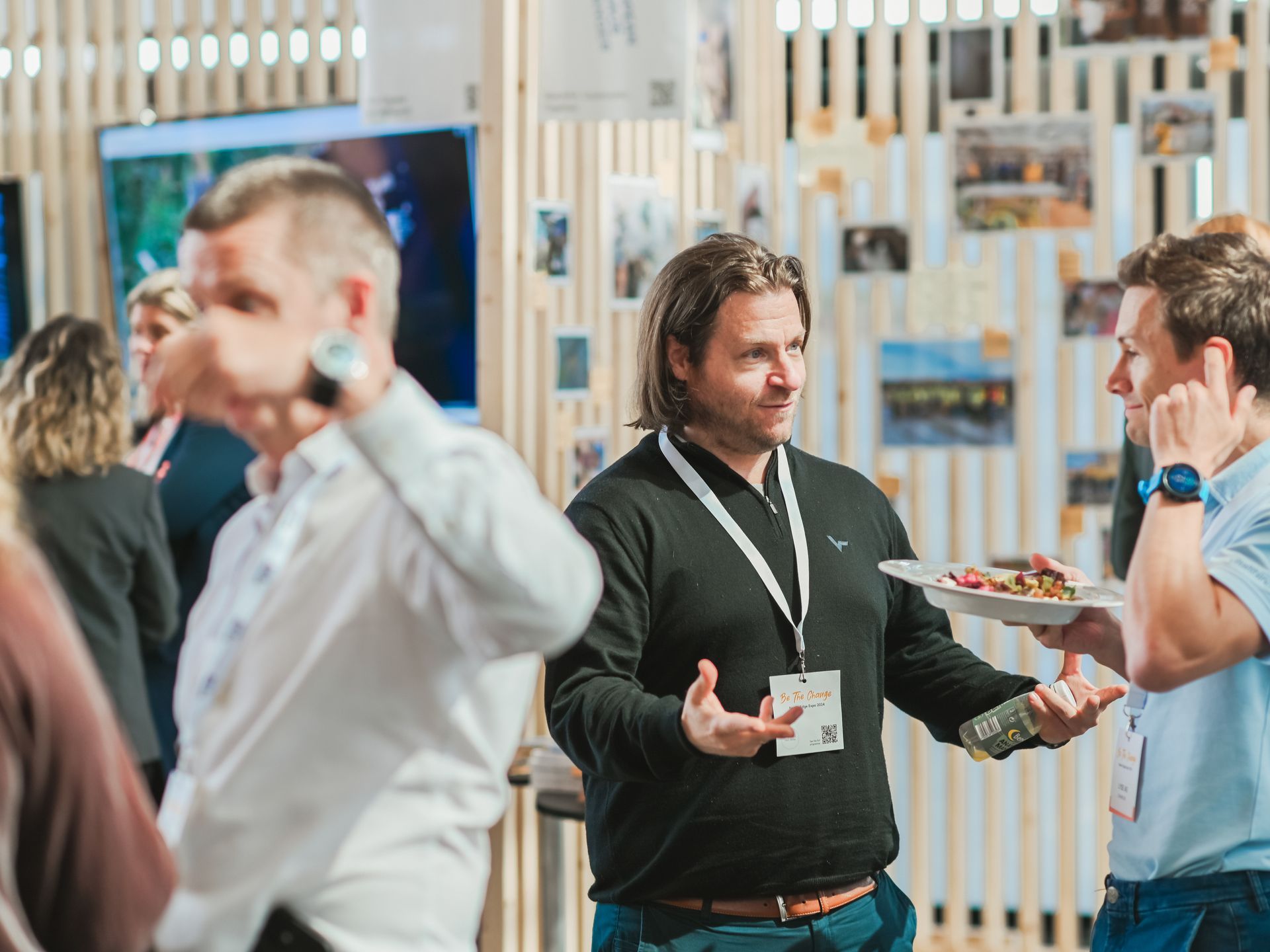 People at a networking event, talking, eating snacks, and wearing name badges. Bright, wooden backdrop.