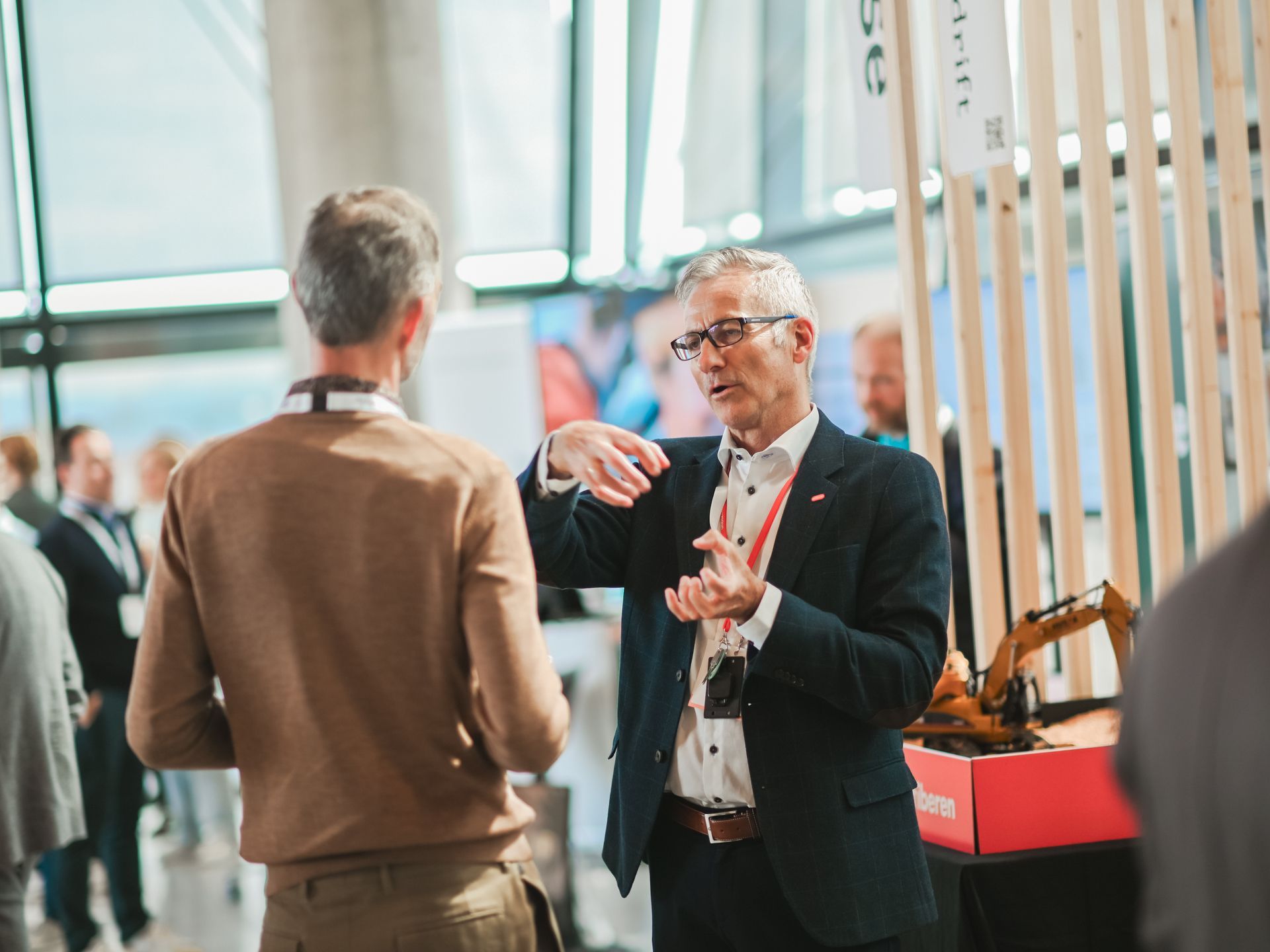 Two men conversing at a trade show. One gestures with hands; others stand nearby.