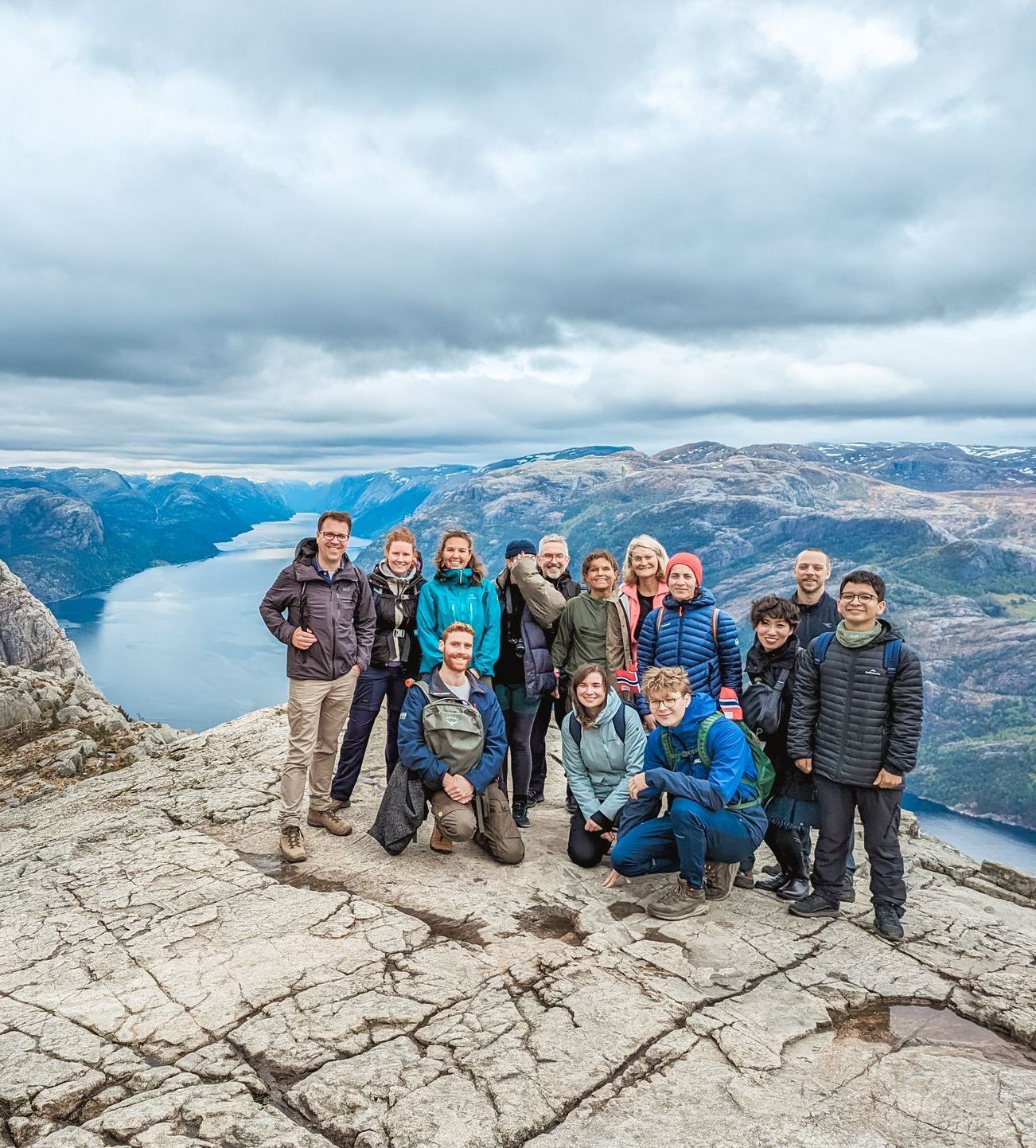 Group of hikers on rocky mountaintop overlooking fjord under cloudy sky.