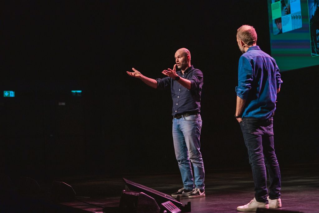 Two men on a stage. One gestures with arms outstretched, the other stands beside him, listening. Dark background.