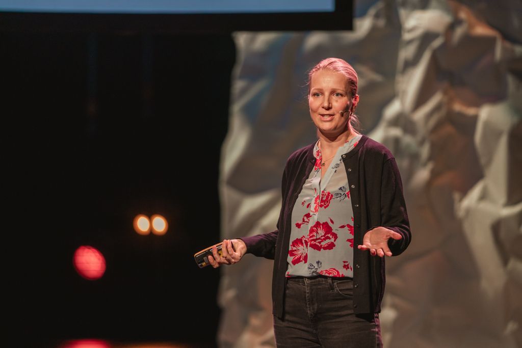 Woman presenting on stage, arms open, holding a clicker. Floral shirt, black cardigan, dark jeans. Bright lights, textured background.