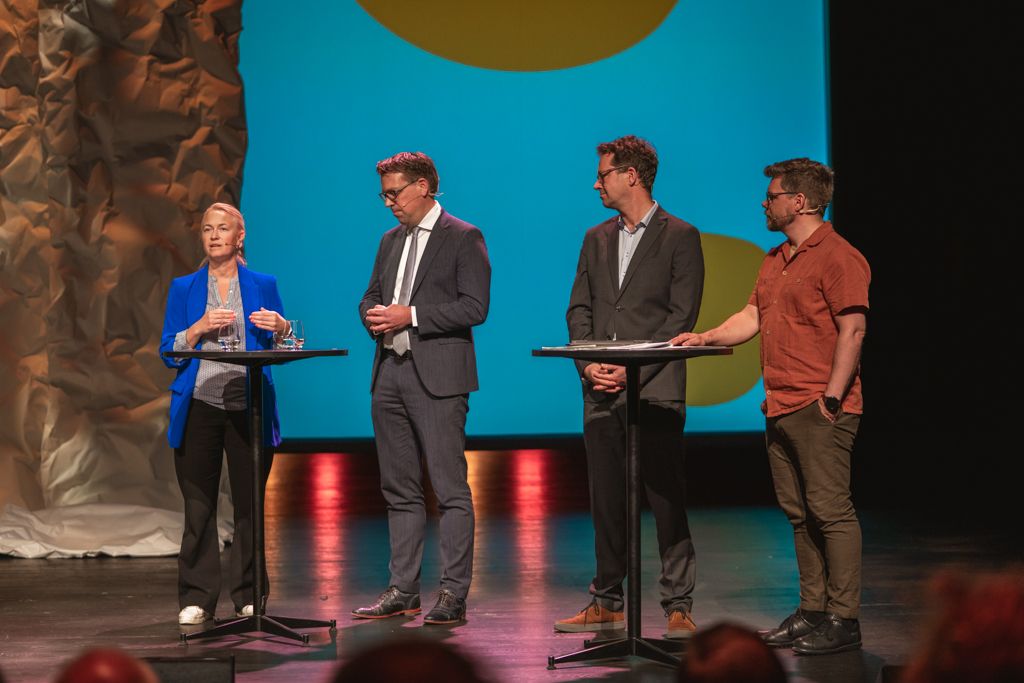 Four people on a stage with podiums. A woman in a blue blazer speaks, others listen. Blue and yellow backdrop.