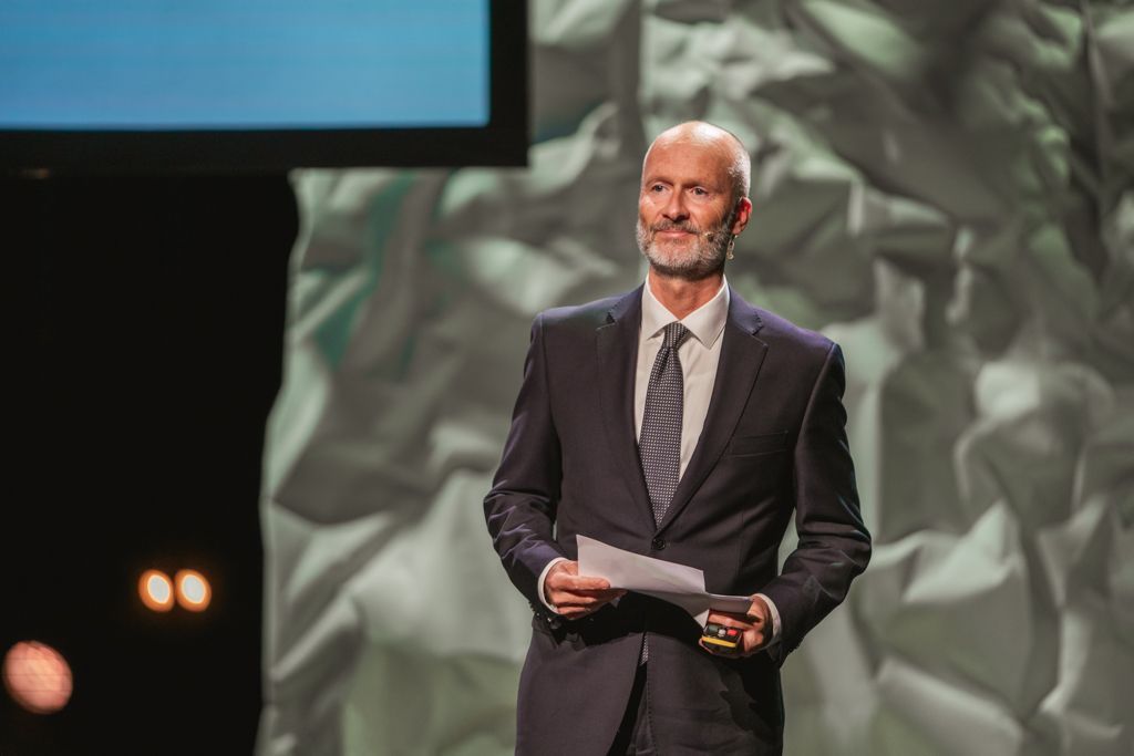 Man in a suit holding papers, speaking on stage with a crumpled paper backdrop and spotlights.