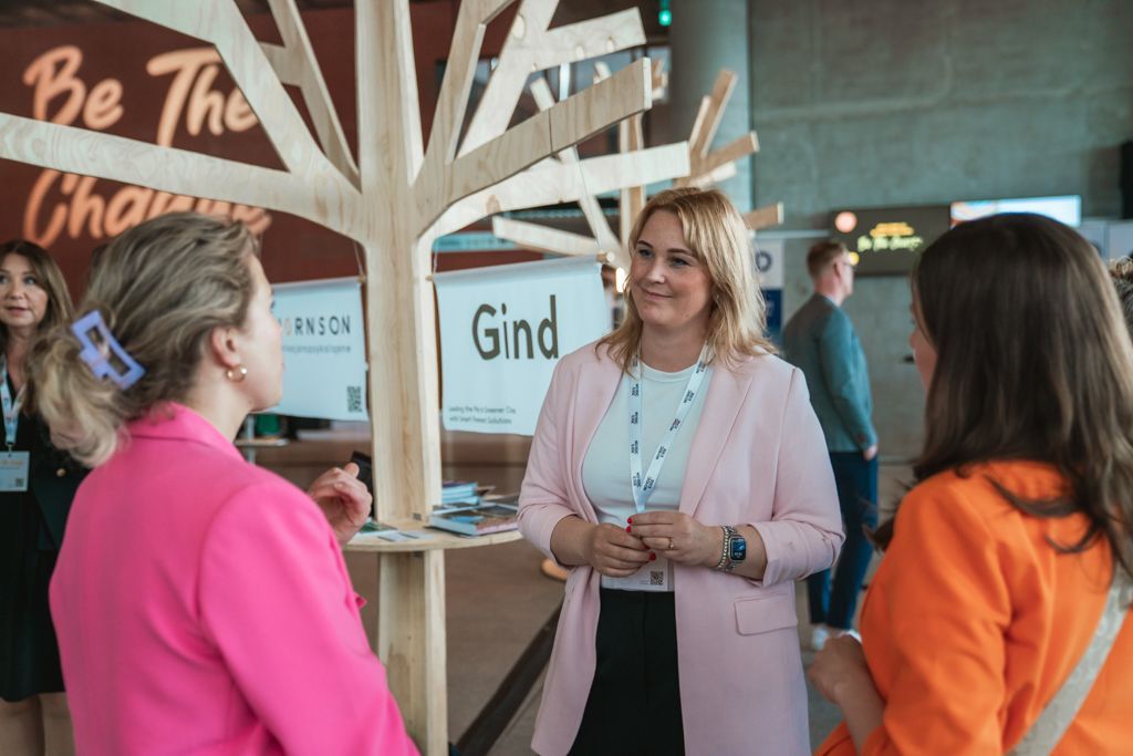 Three women converse near a wooden tree sculpture at a conference. The woman in the center wears a pink blazer and smiles.