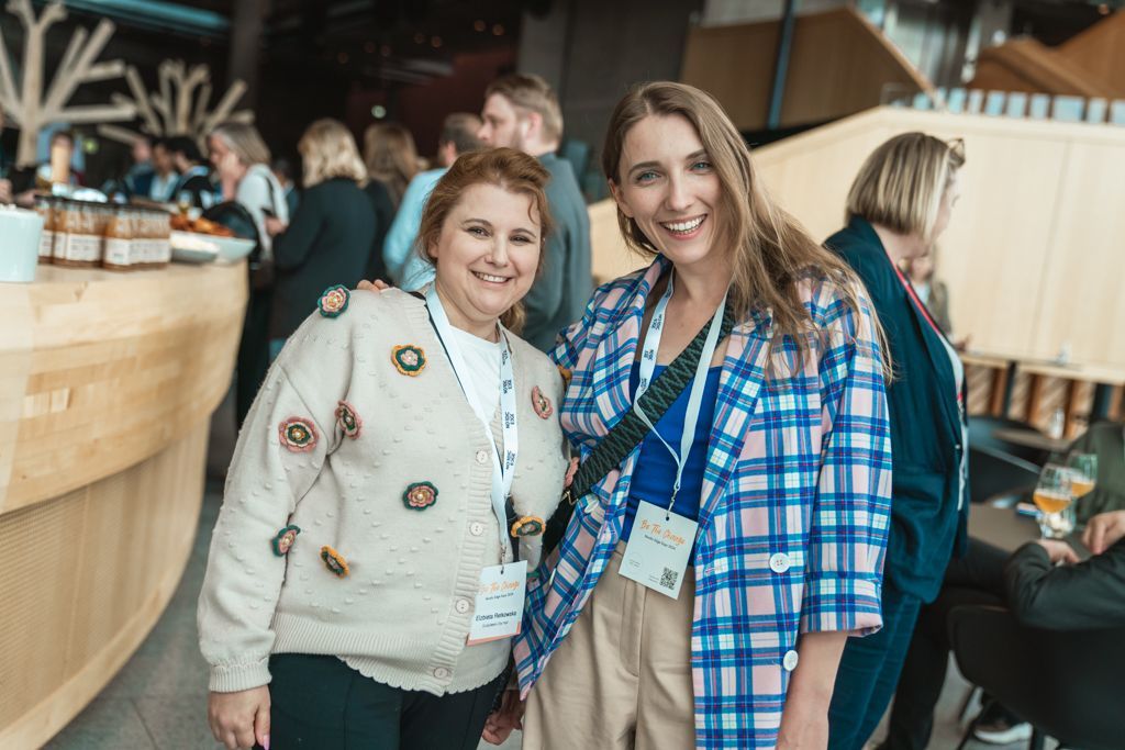 Two women smiling, at an event. One in a plaid jacket, the other in a decorated sweater. Interior shot with a buffet in the background.