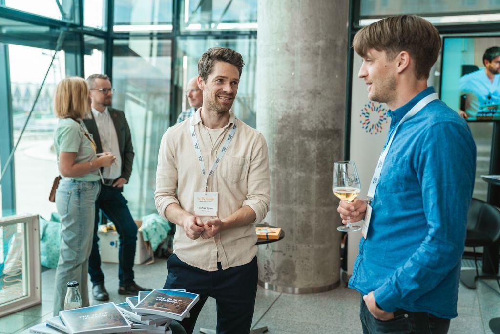 Two men in blue and tan shirts converse, one holding a glass of wine. Others mingle at an event.