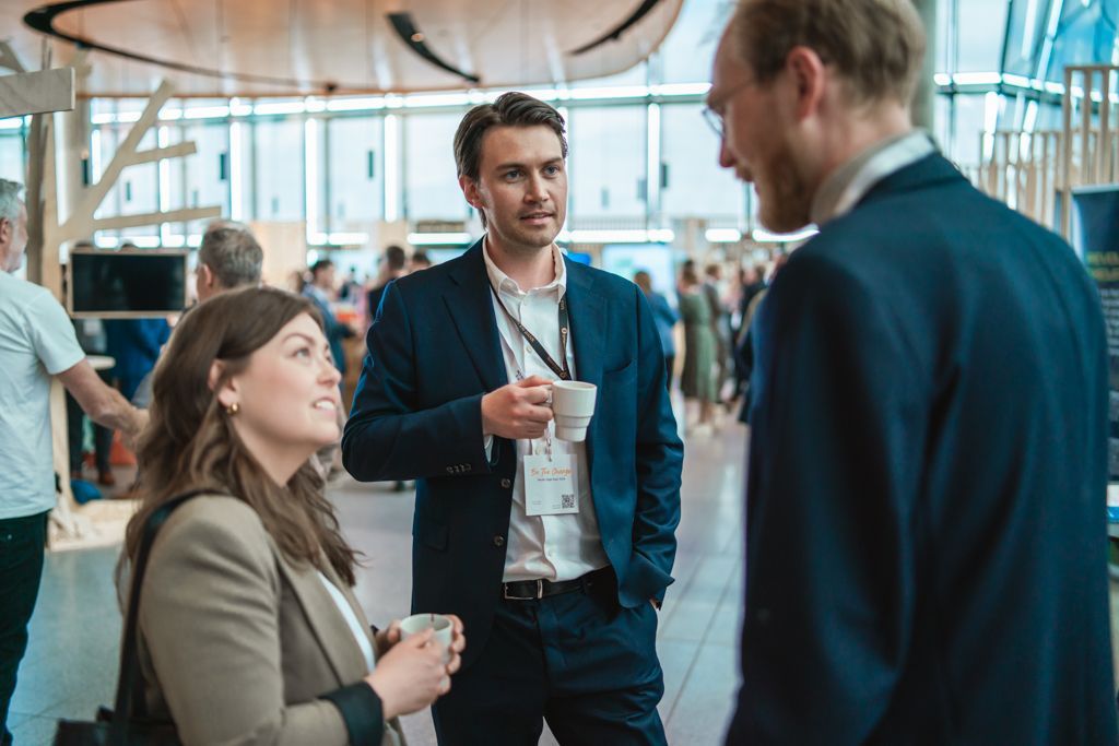 Three people in suits talking at an event, holding coffee cups. Bright, modern interior.