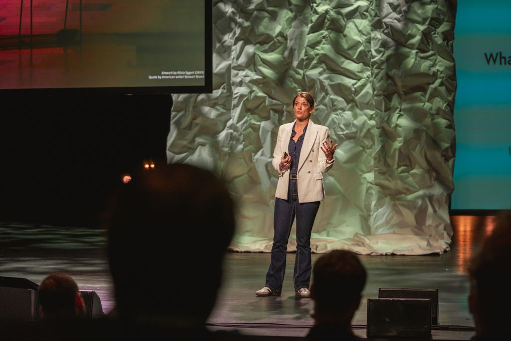 Woman speaking on stage, gesturing, in front of textured backdrop, and audience silhouettes.